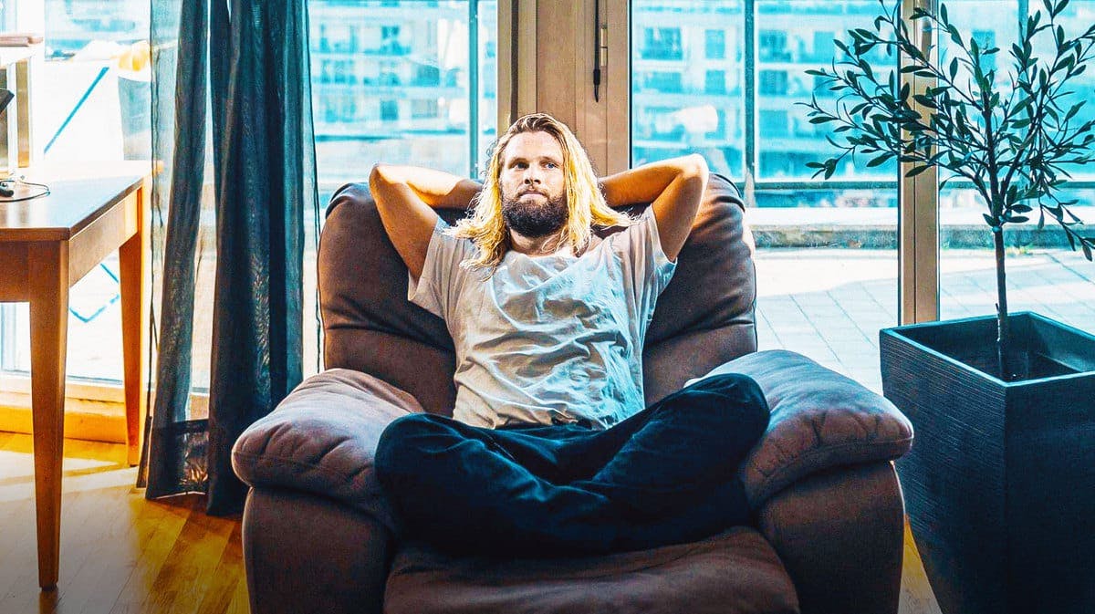 Former Patriots, Michigan football LB Chase Winovich inside a house wearing casual clothes and sitting in a reclining chair, holding a television remote control