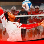 A player on the Nebraska volleyball team spiking over a net with the Nebraska football team in the background