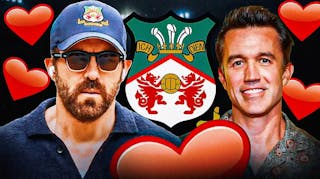 Ryan Reynolds & Rob McElhenney laughing in front of the Wrexham logo, heart emojis in the air