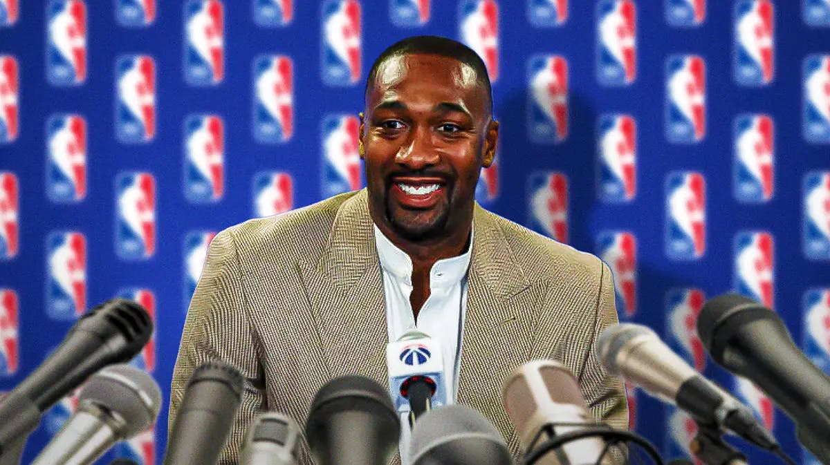 Former player Gilbert Arenas in front of an NBA banner with microphones in front of him.